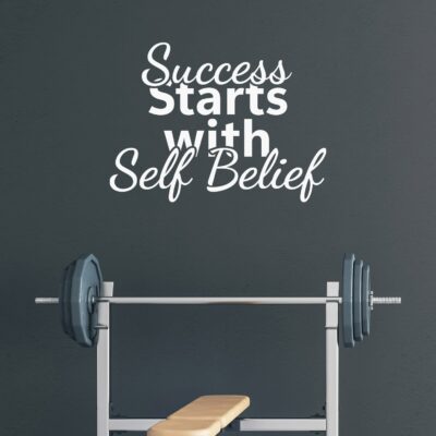 Scritta adesiva motivazionale "Success Starts with Self Belief" su muro grigio in palestra domestica con panca e bilanciere.