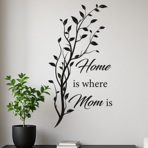 Adesivo murale nero con scritta "Home is where Mom is" su parete chiara, accanto a una pianta in vaso nero in stile minimalista.